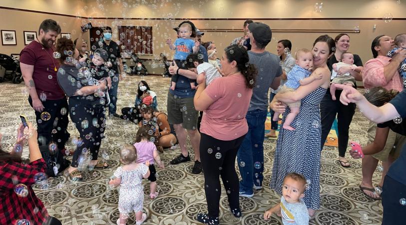 A group of adults and babies are gathered indoors, with many bubbles floating around them.