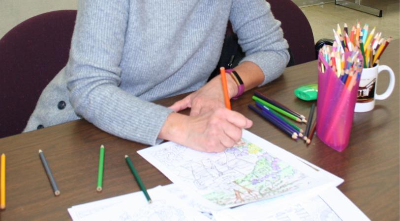 A person is coloring a picture with colored pencils on a wooden table. There are many colored pencils in a pink holder and a white mug.