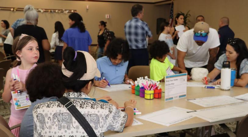 Children and adults sit around classroom tables with craft supplies like colored markers and cups, filling out worksheets while others stand and talk in the background.