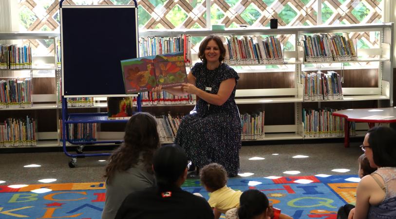 A smiling Ms. Corinne stands in a library reading area, holding a book toward a seated group of children on a colorful floor mat, with shelves of books and a display case in the background.