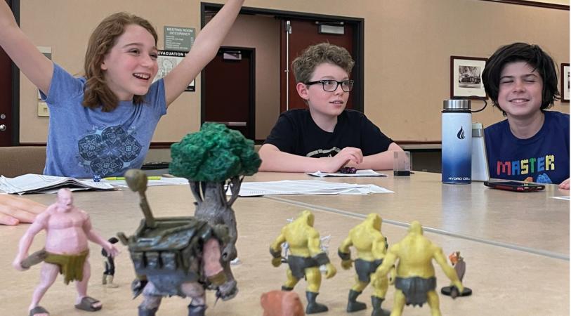 Three young teens are gathered around a table playing a tabletop role-playing game with miniature figures and dice in the foreground.