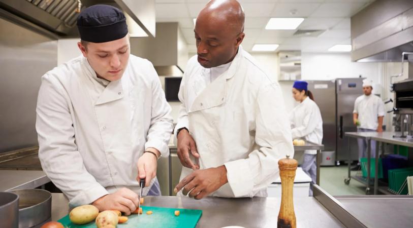 Two chefs in white coats work at a stainless-steel prep table, cutting vegetables on a blue cutting board with onions nearby, while others cook in the background in a professional kitchen.