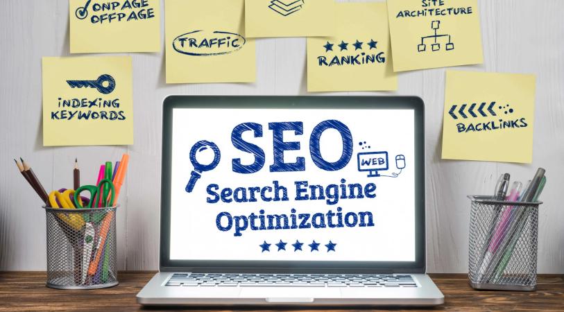 Laptop displaying “SEO Search Engine Optimization” with star ratings, surrounded by sticky notes labeled content, traffic, ranking, site architecture, indexing keywords, and backlinks, with pencils in desk holders.