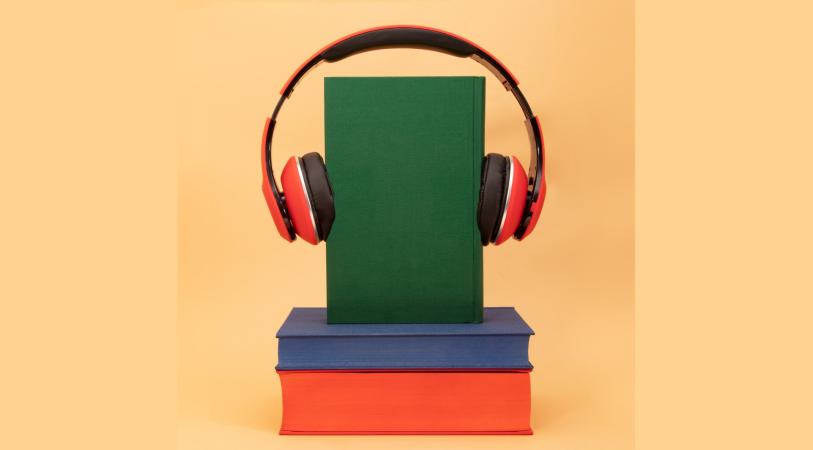 A green book stands on top of a stack of two books, one blue and one orange. A pair of red headphones rests over the green book.